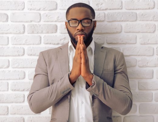 Handsome Afro American man in classic suit and glasses is keeping palms together like praying, standing against white brick wall